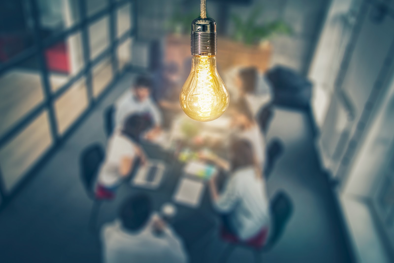 An artistic photo of a lightbulb in front of a group of people meeting at at conference table